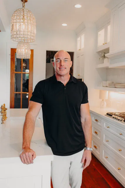 Portrait of a smiling man with a shaved head wearing a black polo shirt and white pants in a modern white kitchen with gold accents and a chandelier.