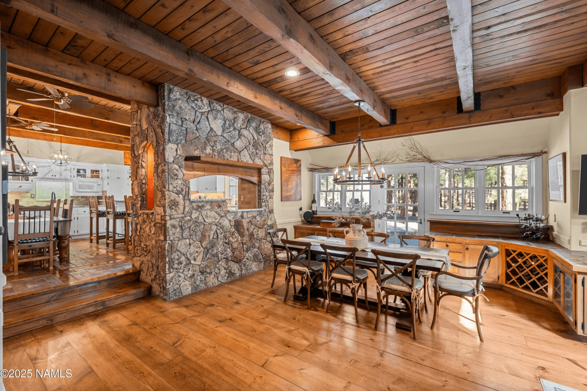 Rustic dining room with wooden floors, stone fireplace, cozy windows, and natural light, connecting to the kitchen.