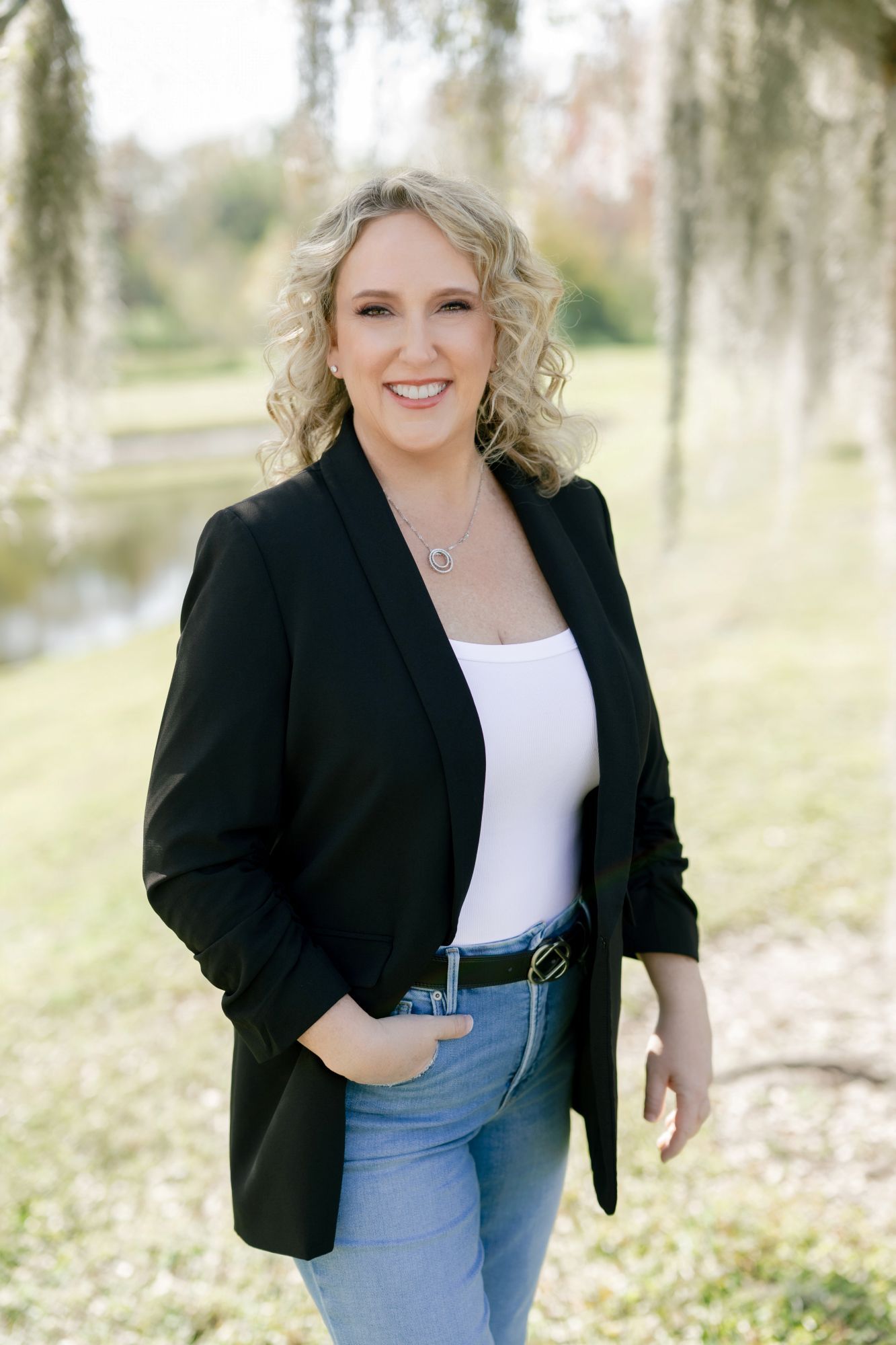 Smiling woman with blonde curly hair wearing black blazer, white tank top, blue jeans, standing outdoors near a pond with trees in the background.