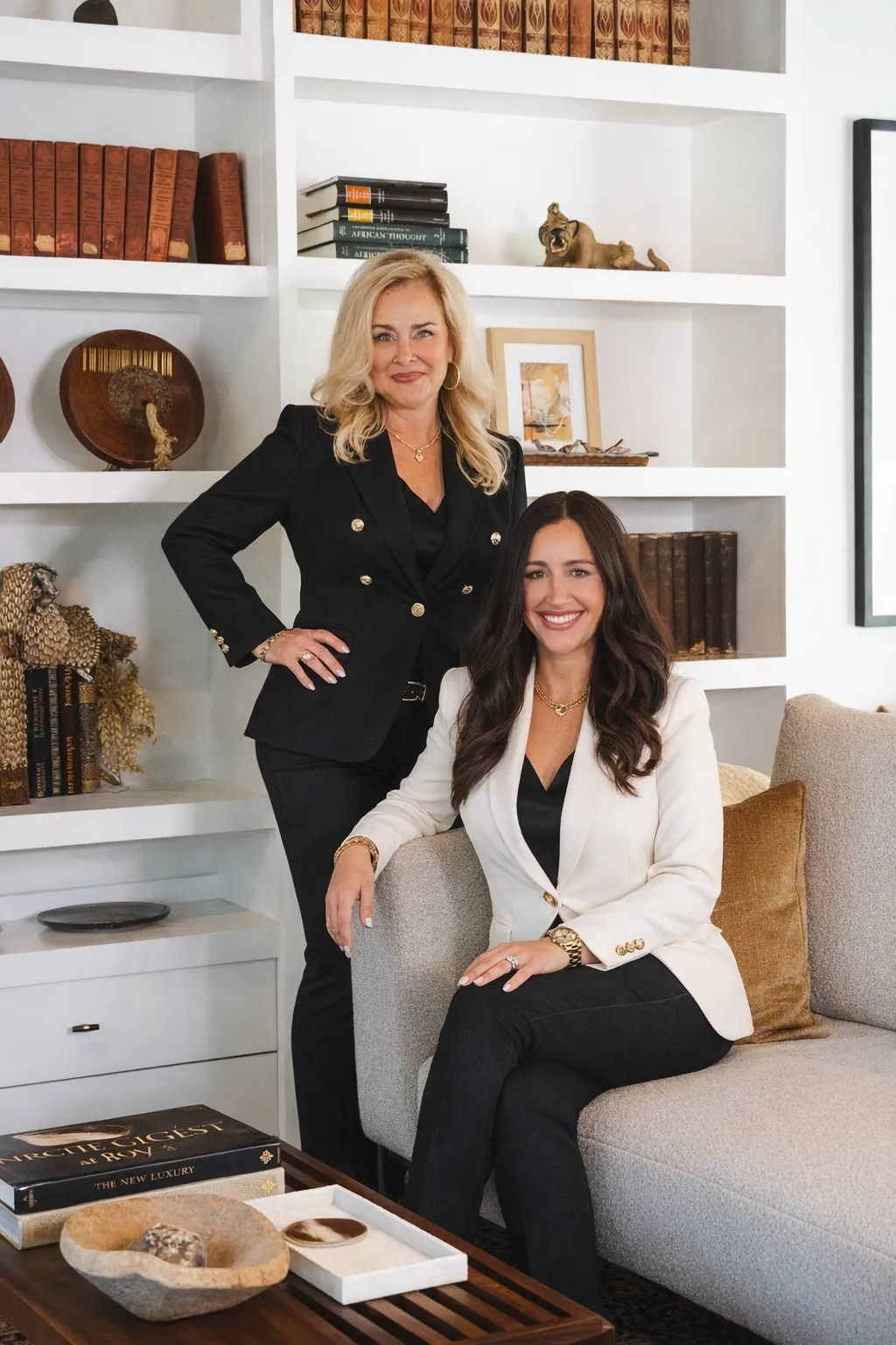 Two women in elegant black outfits posing on a staircase indoors, with one wearing leopard print high heels and the other with a leopard print belt, smiling at the camera.
