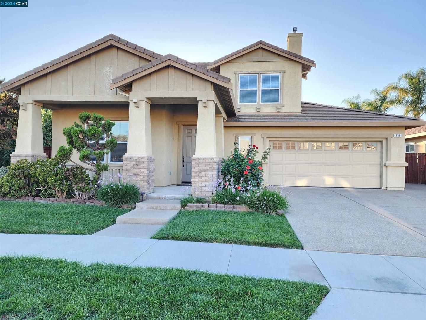 Two-story beige stucco house with a landscaped front yard, featuring a manicured lawn, flowering bushes, and a small tree, with a front porch and a two-car garage.