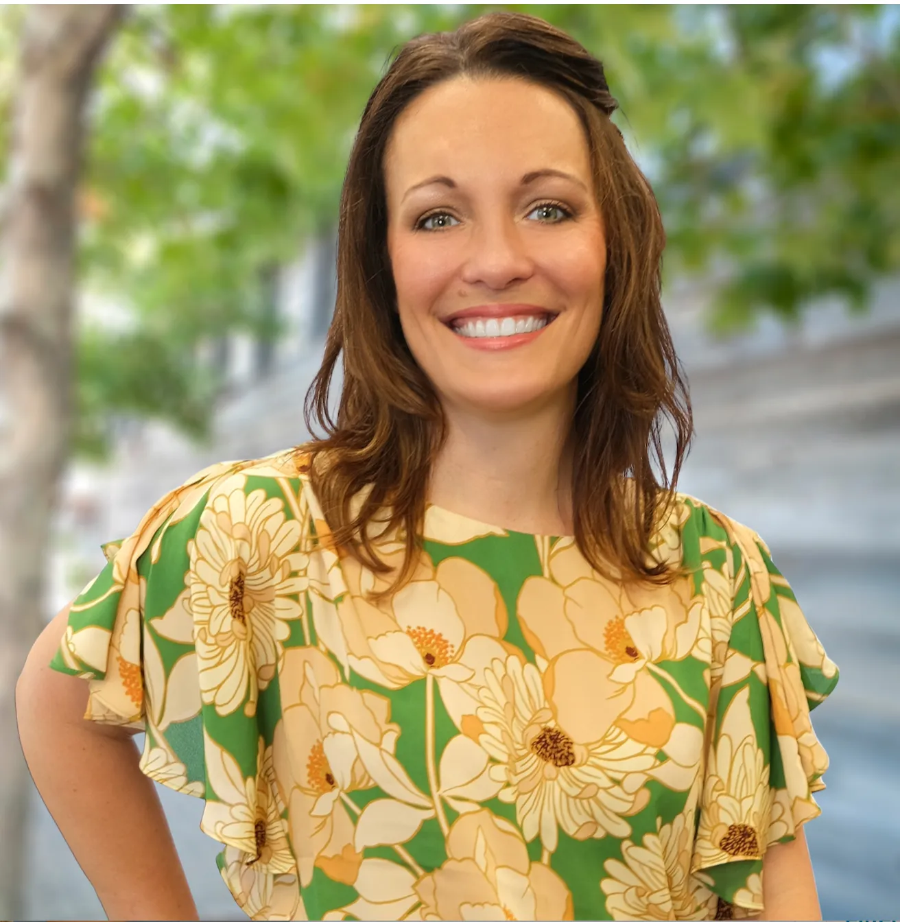 Smiling woman with shoulder-length brown hair wearing a green and yellow floral dress outdoors with green trees in the background.
