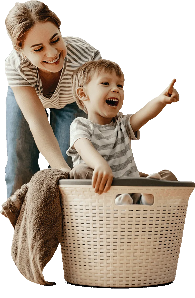 Happy child pointing and smiling while riding in a laundry basket, with a woman smiling beside him, indoor setting, joyful family moment.