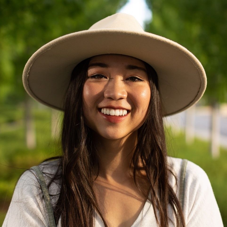 Smiling woman wearing a wide-brimmed hat outdoors in a park with green trees in the background