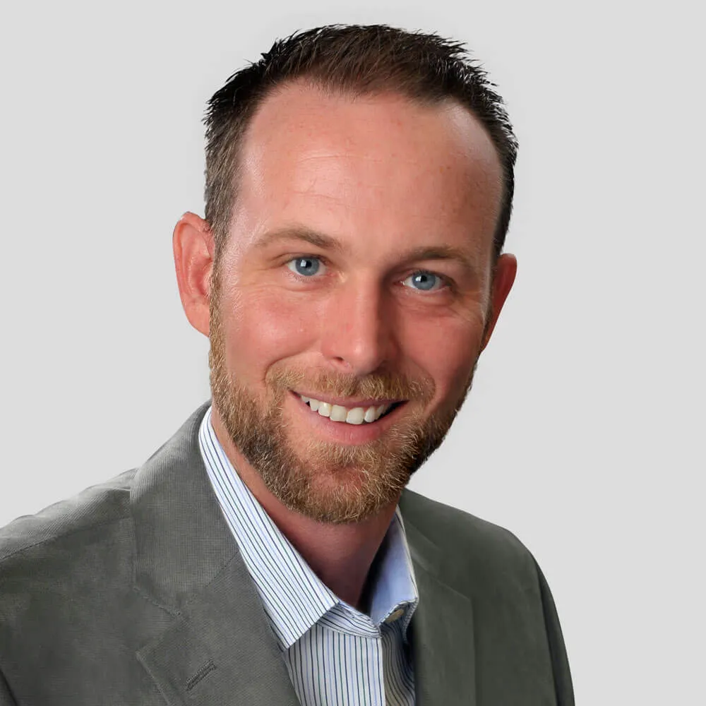 Professional headshot of a smiling man with short dark hair, blue eyes, and beard, wearing a gray suit and blue striped shirt against a plain light gray background.
