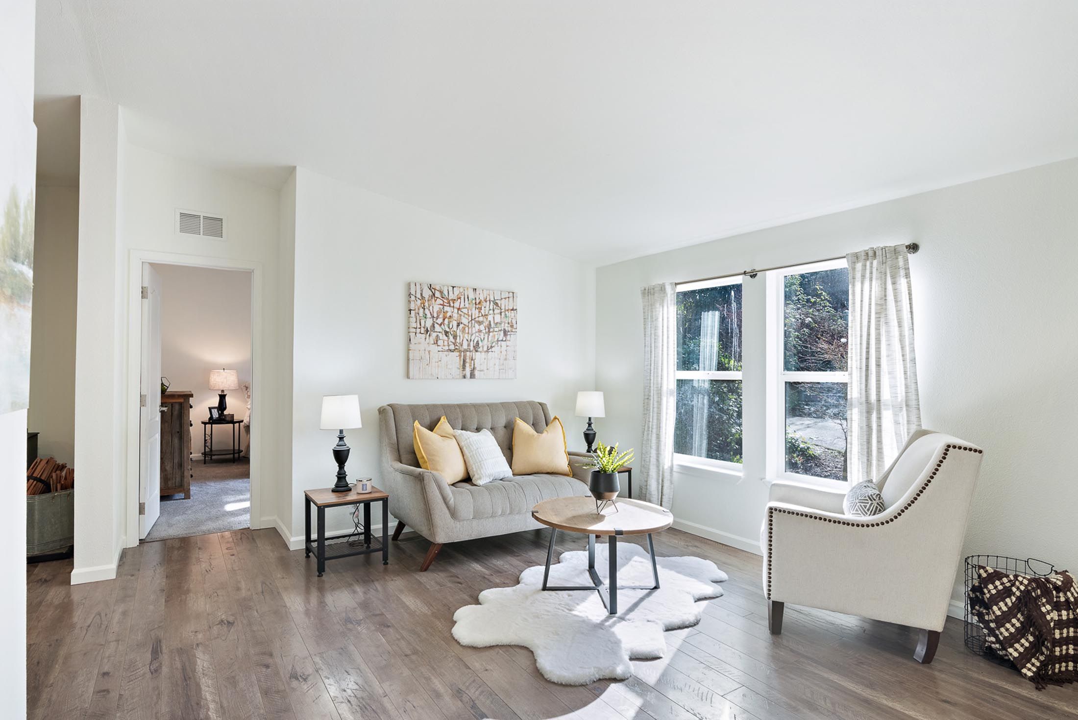 Cozy living room corner with two gray upholstered swivel chairs, a round side table with a white ceramic table lamp, a large green potted plant, and a window showing a garden view with trees and blue sky.