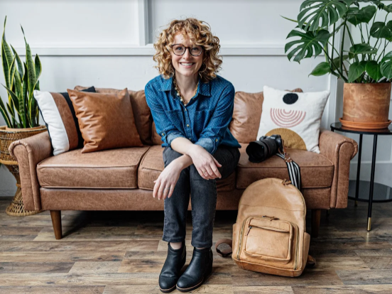 Smiling woman with curly hair and glasses sitting on a brown sofa in a modern living room with green plants, a backpack, and a camera.