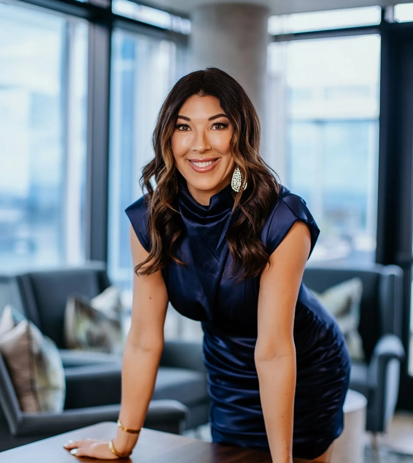 Professional businesswoman with long wavy brown hair, wearing a black blazer, gold necklaces, and light makeup, smiling in a modern office setting.