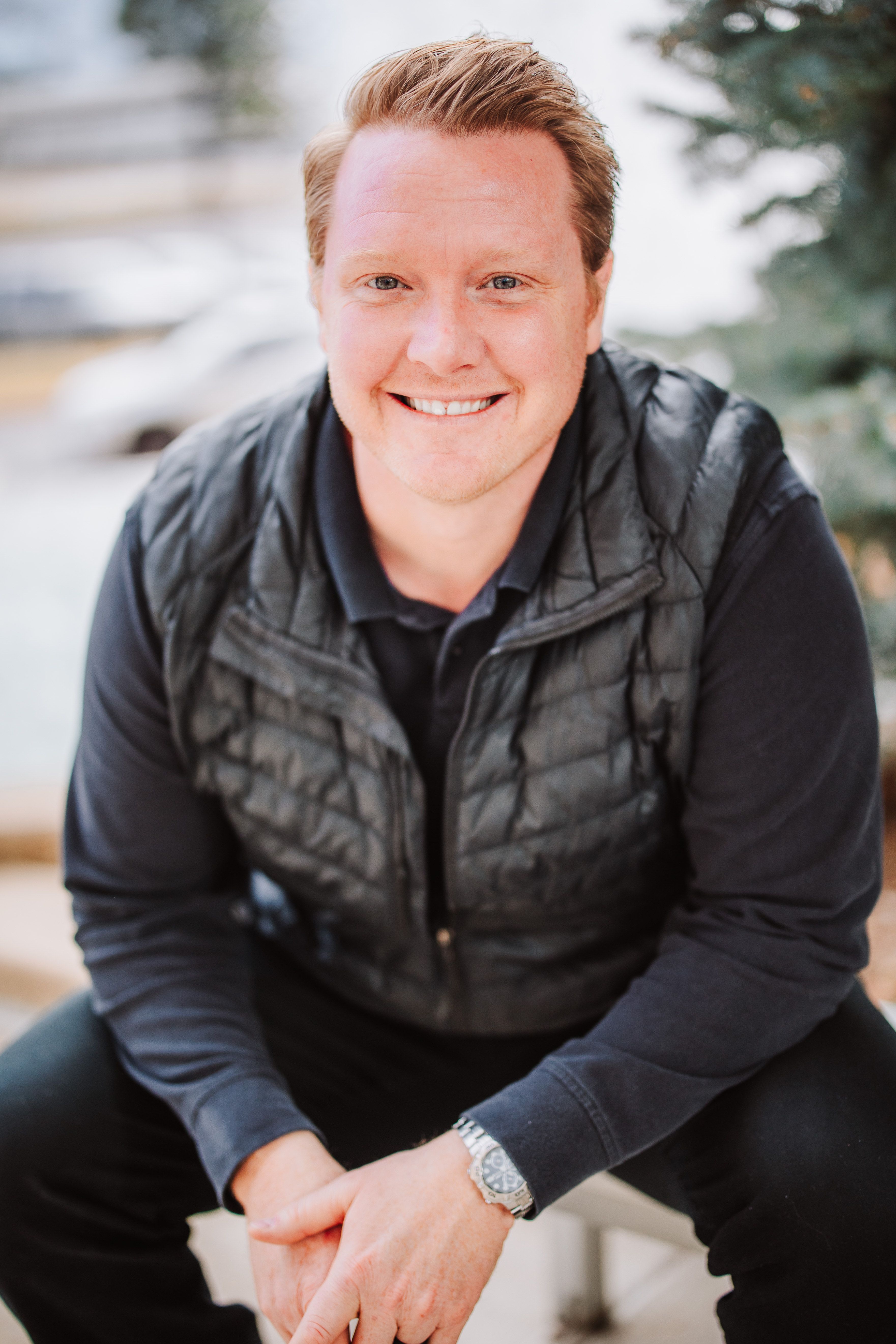 Portrait of a smiling man with red hair, wearing a black quilted vest and black long-sleeve shirt, sitting outdoors with blurred background of trees and cars.