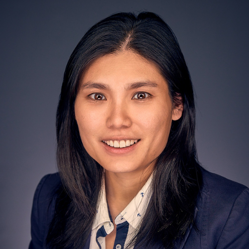 Professional portrait of a young Asian woman with long black hair wearing a navy blazer and white shirt, smiling against a dark gray background.