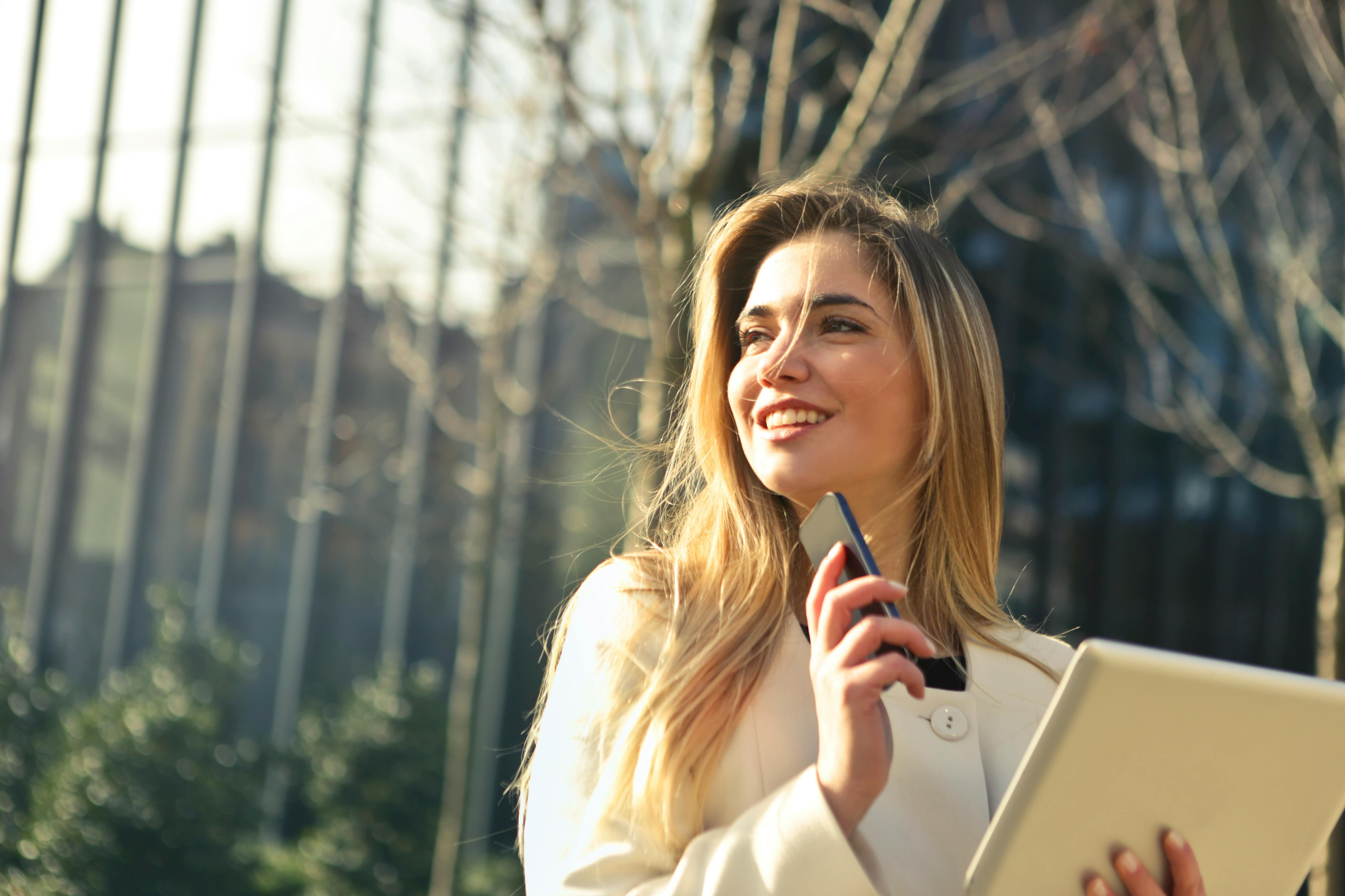 Young woman holding a smartphone and a folder outdoors near modern glass buildings, smiling and enjoying the sunny day.
