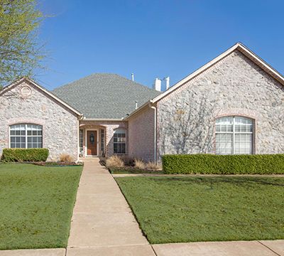 Single-story brick house with a manicured lawn, concrete pathway, and large front windows under a clear blue sky