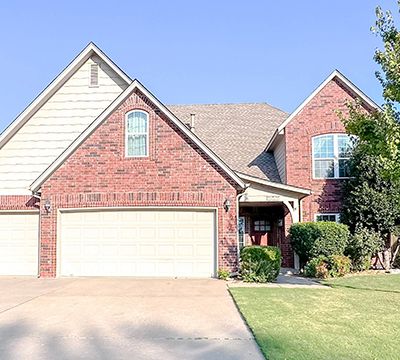 Two-story suburban house with a brick and siding exterior, attached two-car garage, well-maintained front lawn, and landscaping under a clear blue sky.