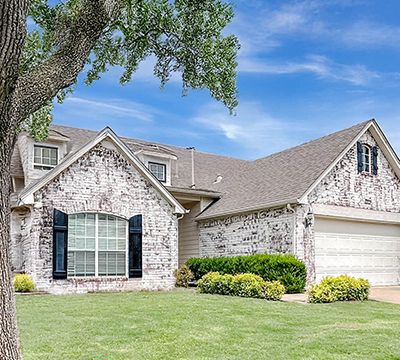 Beautiful suburban house with brick and stone exterior, large front window with black shutters, well-maintained lawn, and a two-car garage under a blue sky