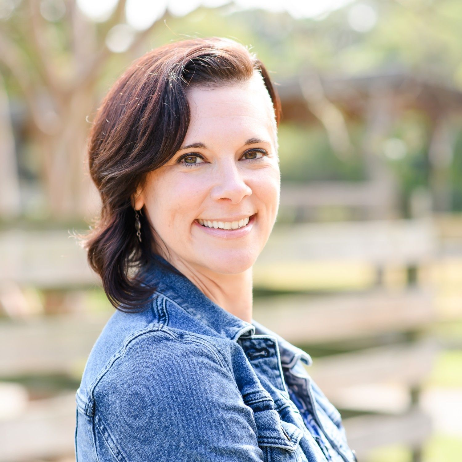 Smiling woman with short dark hair wearing a denim jacket outdoors in a park during daytime