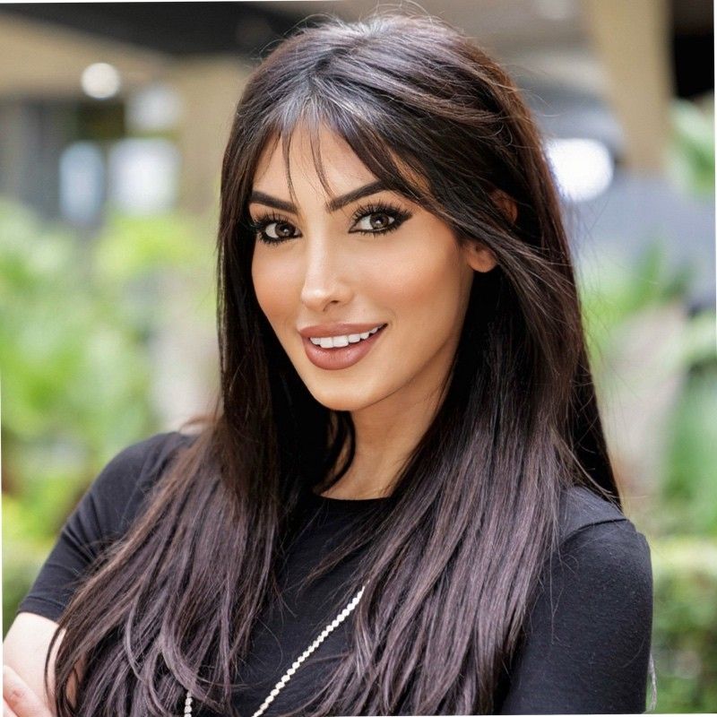 Smiling woman with long dark hair and makeup, wearing a black top, posed outdoors with blurred greenery background