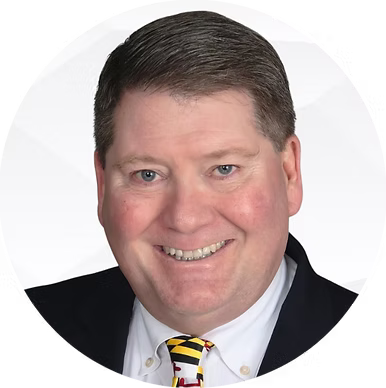 Headshot of a middle-aged man with short dark hair, wearing a dark suit, white shirt, and a yellow tie with black and red stripes, smiling against a white background.