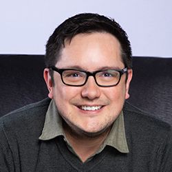 Smiling man with dark hair glasses and casual clothing sitting against light background