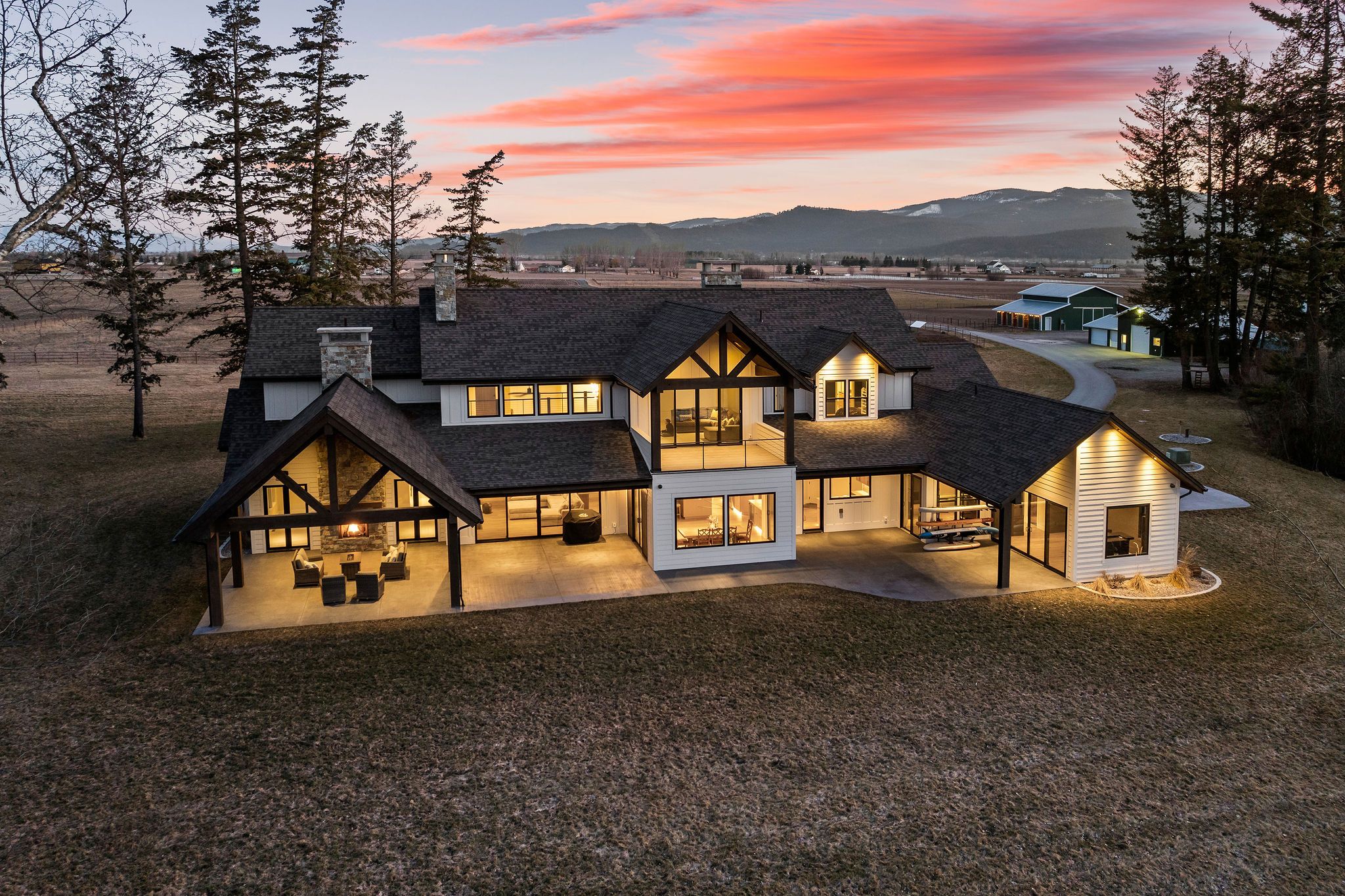 Elegant living room with a stone fireplace, large flat-screen TV mounted on gray paneled wall, plush beige sofas, tufted ottoman, large window with curtains, and decorative items on built-in shelves.