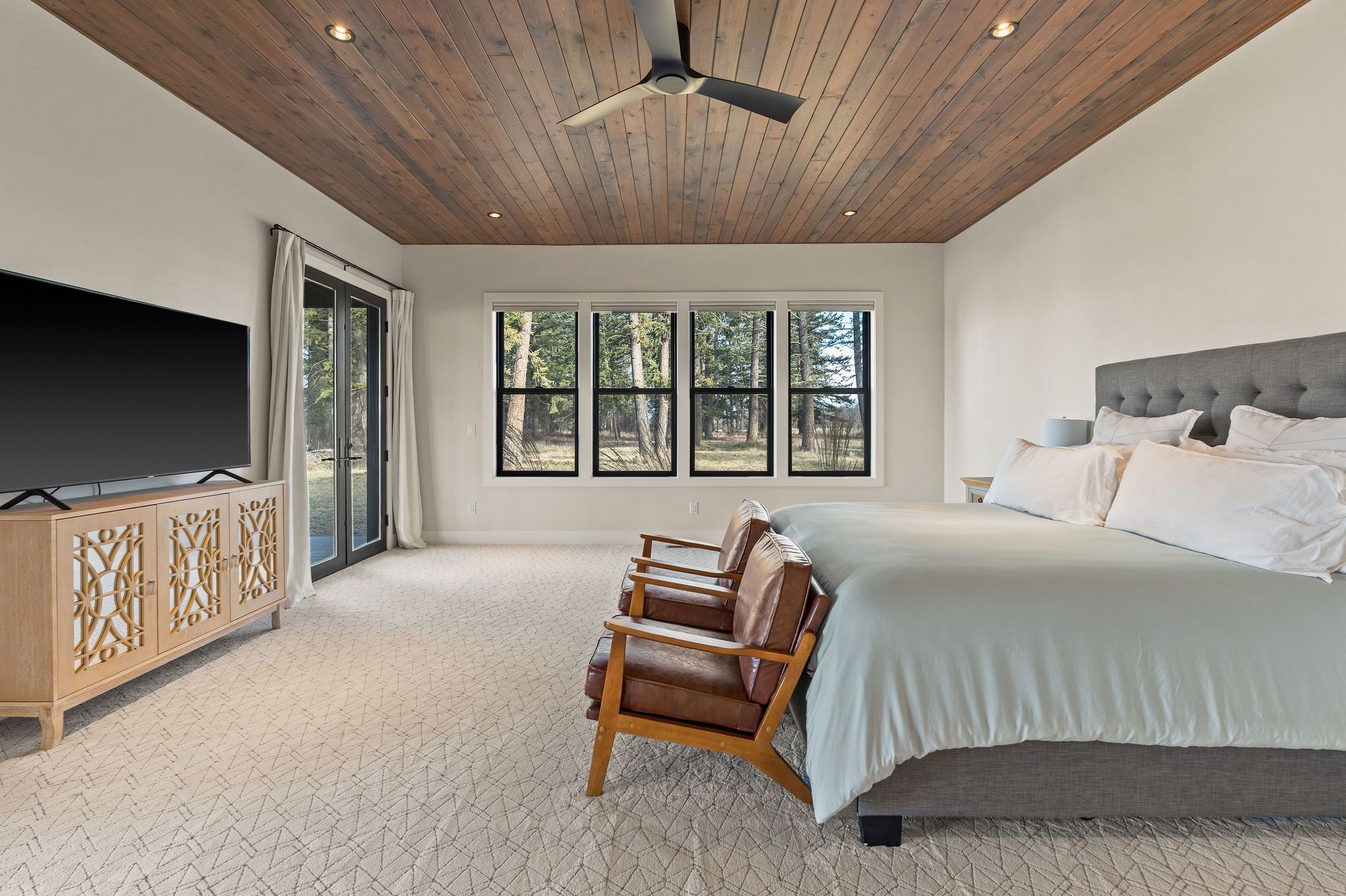Elegant living room with a stone fireplace, large flat-screen TV mounted on gray paneled wall, plush beige sofas, tufted ottoman, large window with curtains, and decorative items on built-in shelves.