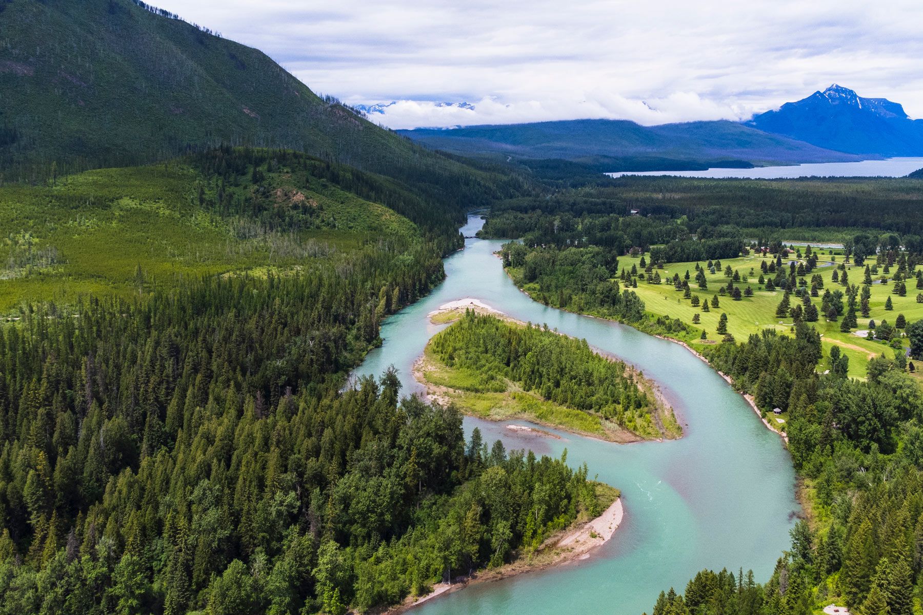 Aerial view of a residential neighborhood surrounded by dense green forest with a large body of water in the background, under a clear blue sky.