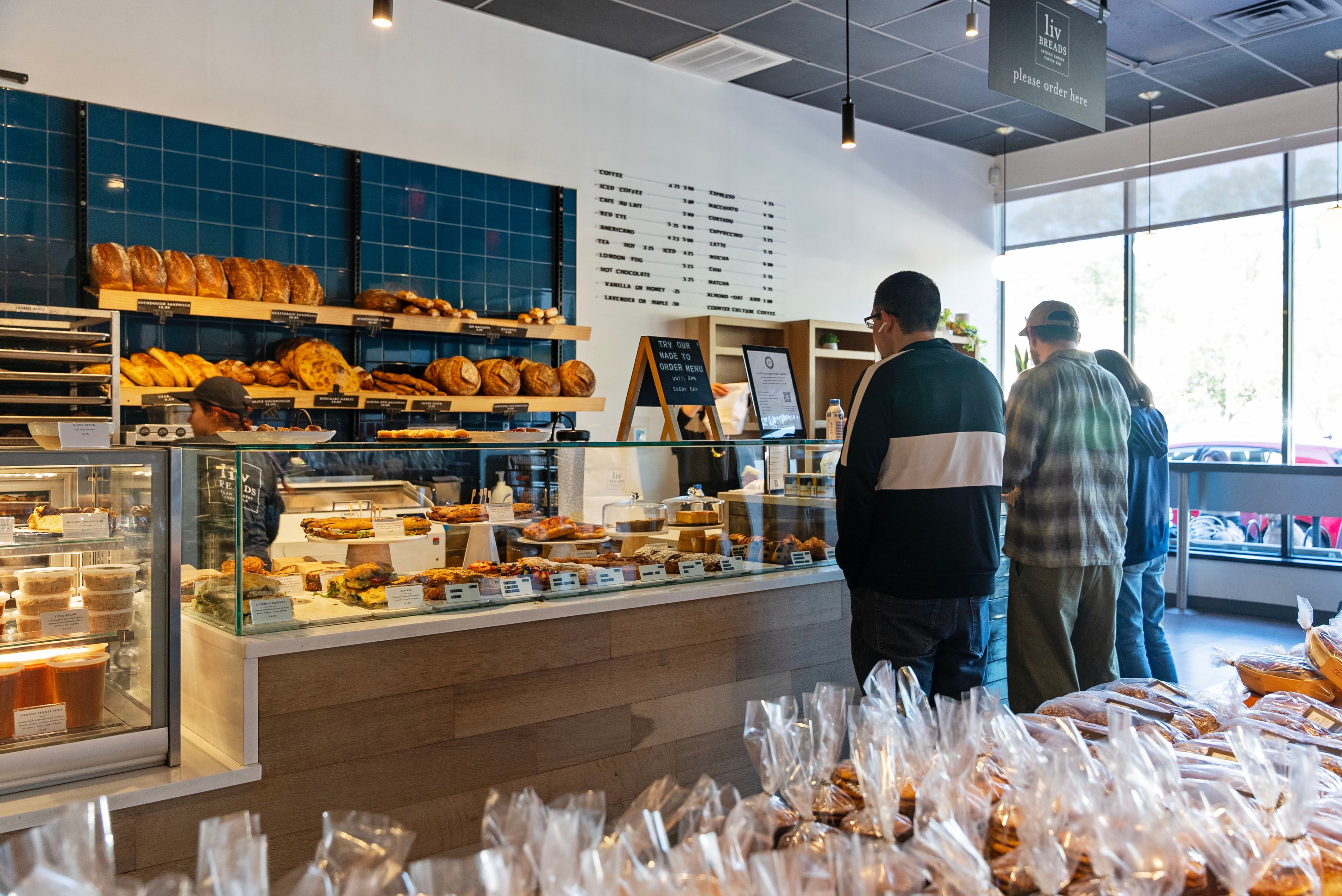 Customers ordering baked goods at a bakery counter with a variety of bread and pastries on display, inside a modern café with large windows and a minimalistic design.
