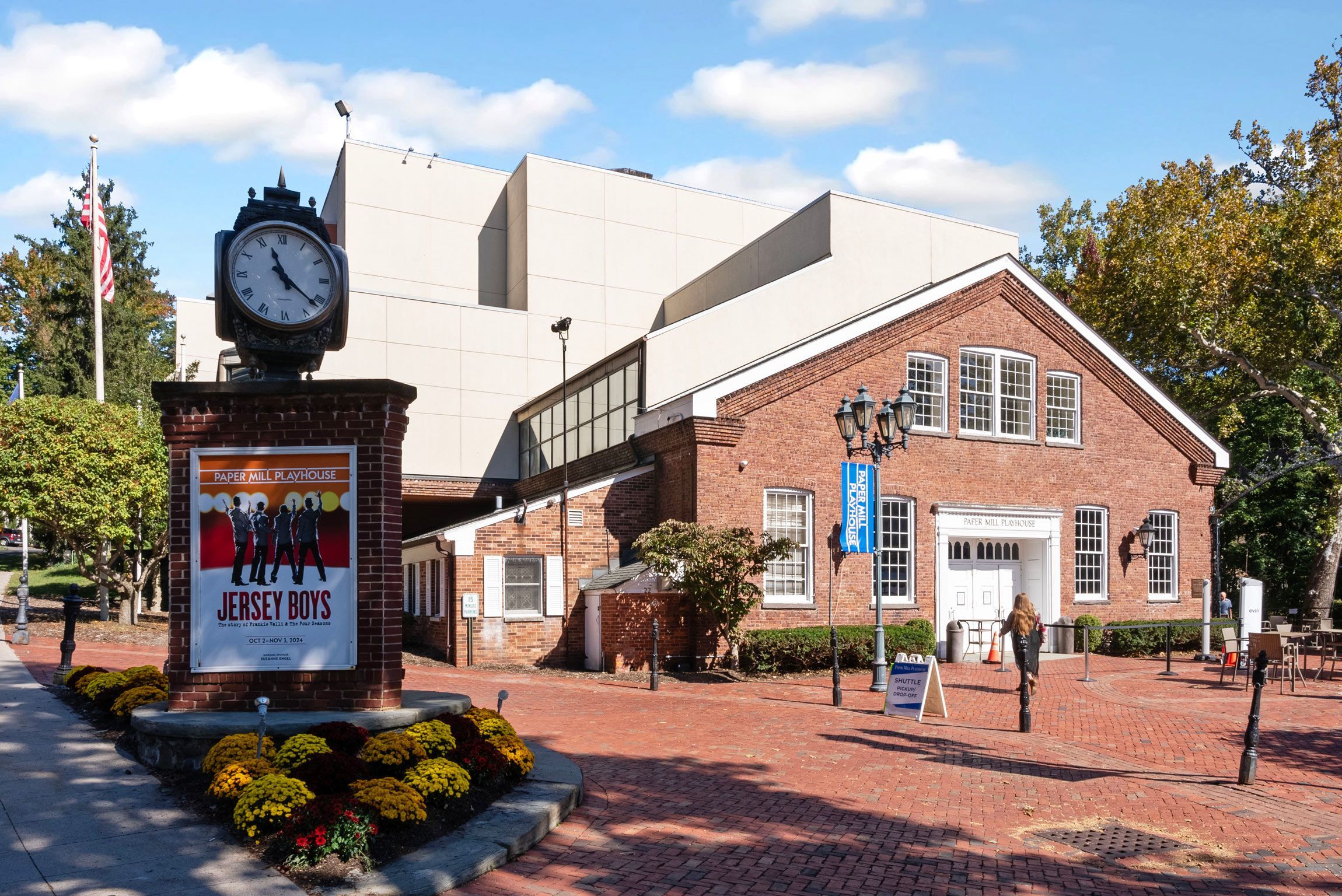 Exterior view of the Paper Mill Playhouse in Millburn, New Jersey, showcasing the brick building, vintage clock, trees, and fall foliage, with promotional banners for 