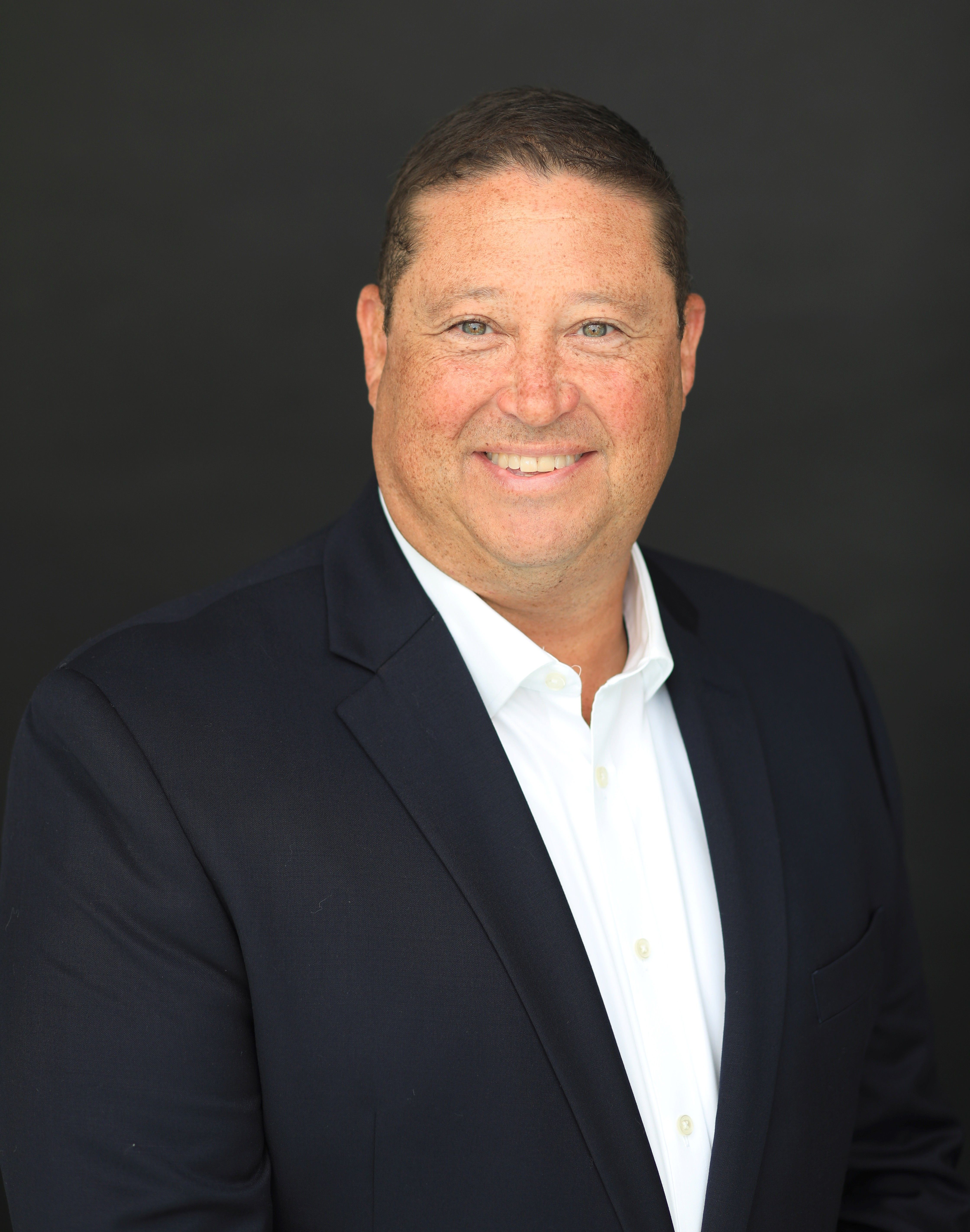 Portrait of a young woman with long wavy brown hair and blue eyes, wearing a white top, smiling softly, with green indoor plants in the background.