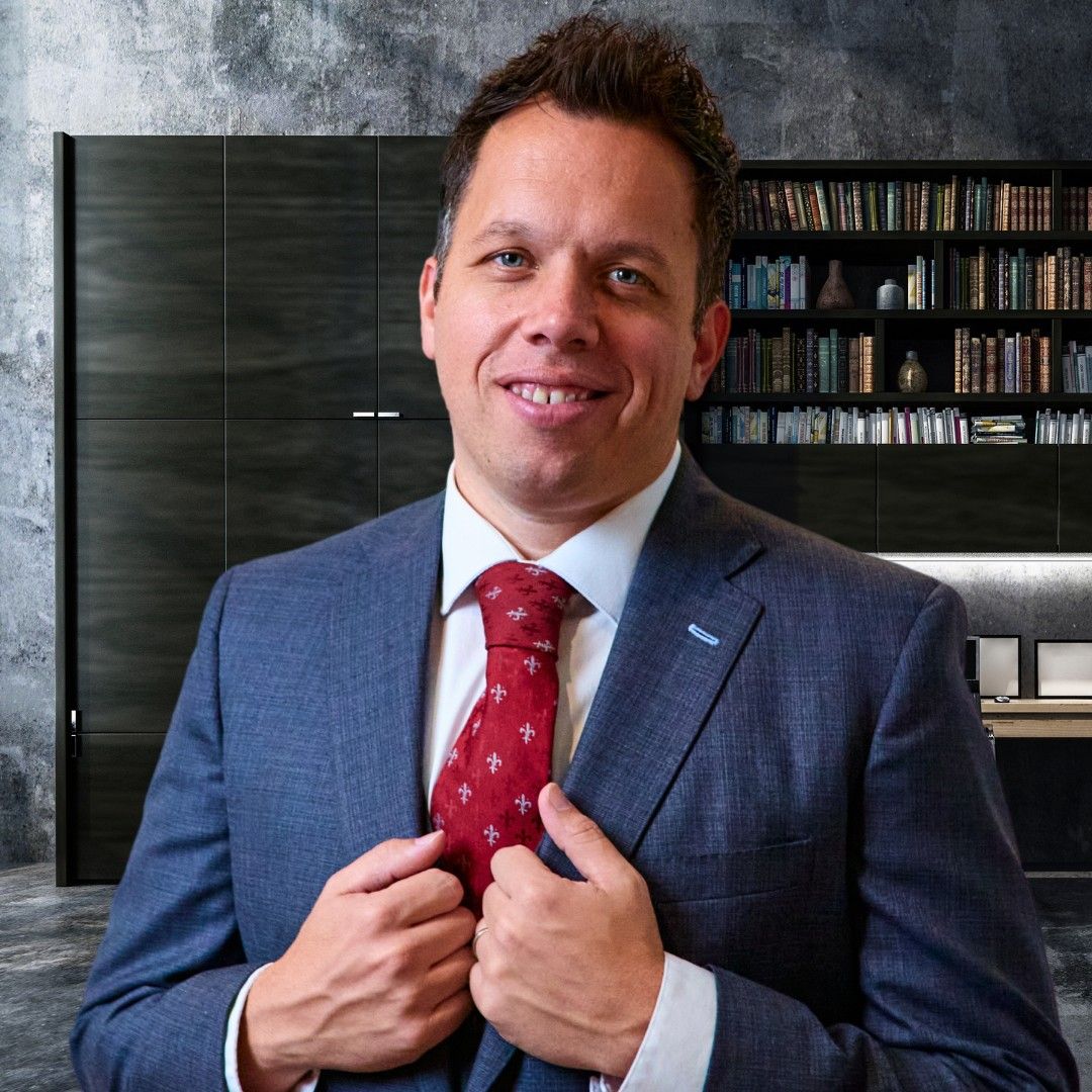 Smiling elderly man with gray hair, glasses, pink shirt, and maroon sweater in a modern office setting.