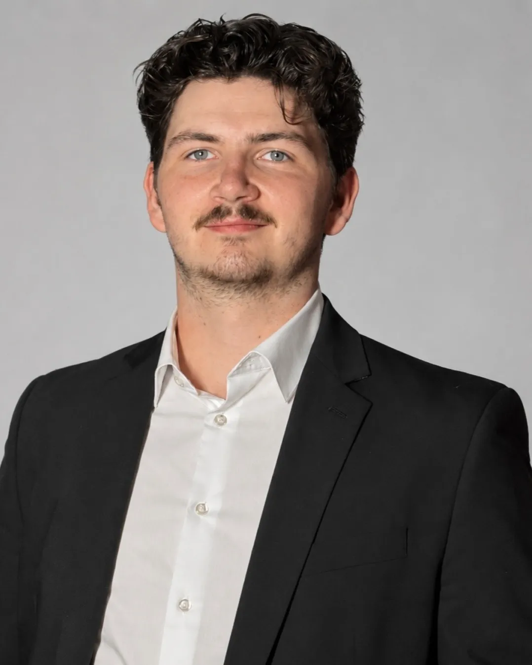 Professional headshot of a young man with curly dark brown hair, light blue eyes, and a mustache with light stubble, wearing a black blazer over a white button-up shirt, photographed against a light gray background.
