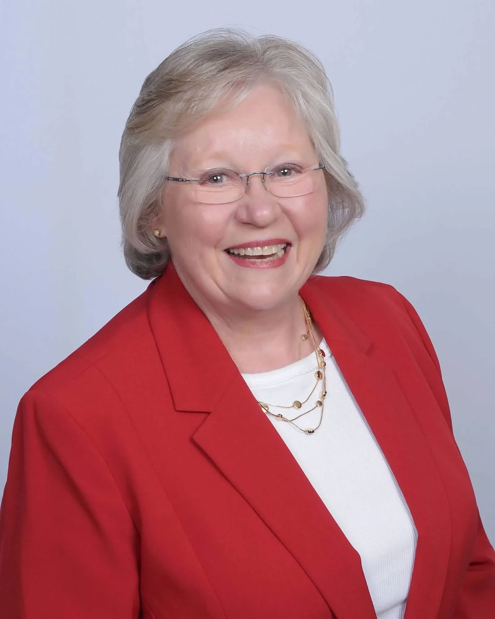 Professional headshot of a smiling older woman with short, wavy blonde-gray hair and rimless glasses, wearing a bright red blazer over a white top with a layered gold necklace, photographed against a light gray background.