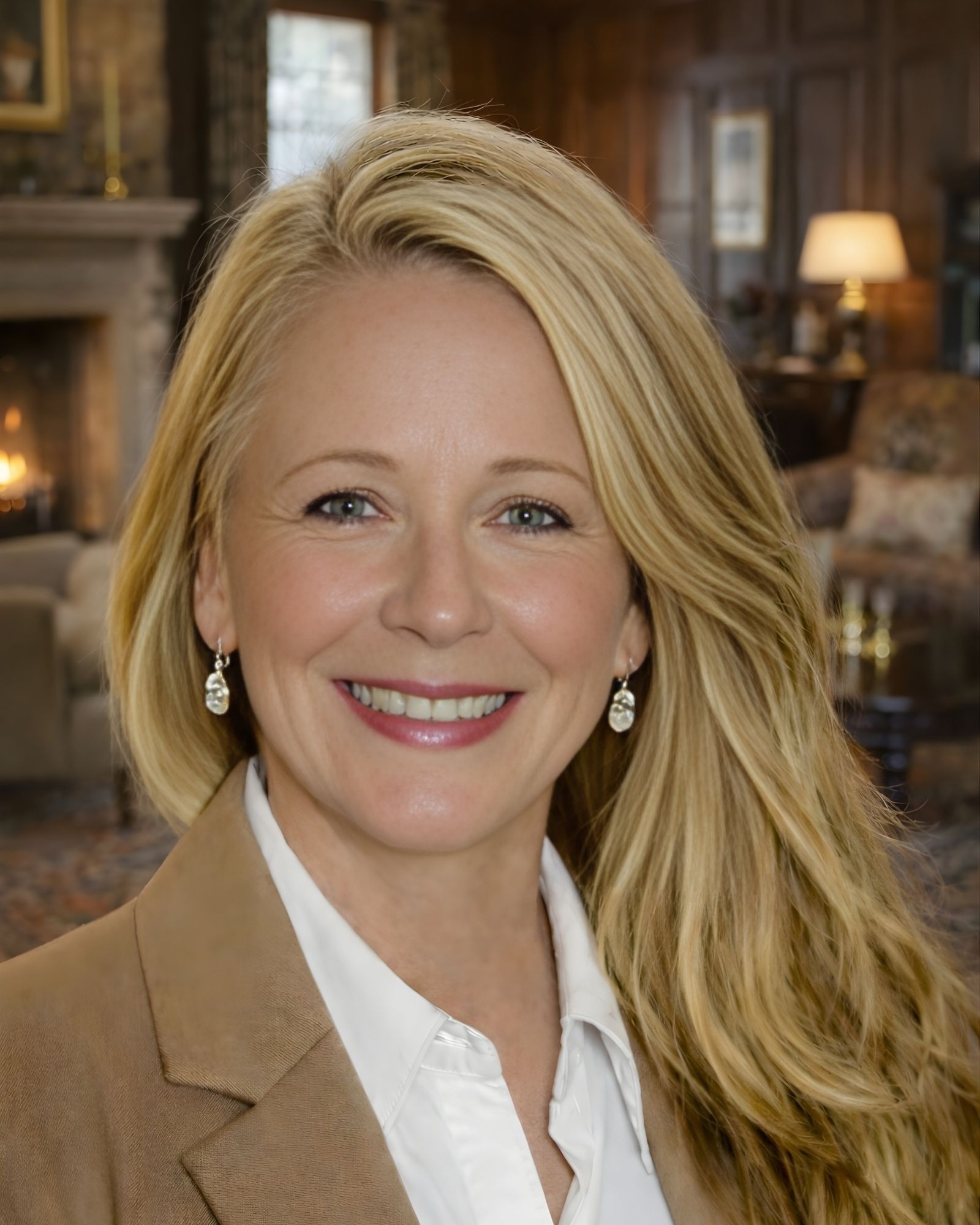 Portrait of a smiling woman with long auburn hair, wearing a black blazer and white shirt, standing outdoors with autumn foliage in the background