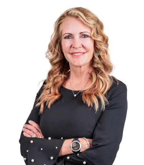 Smiling professional woman with blonde curly hair wearing a black long-sleeve top and accessories, posing with arms crossed against a white background.