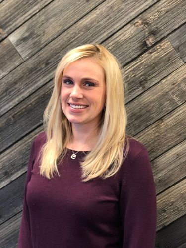 Portrait of a smiling woman with long blonde hair wearing a burgundy sweater standing in front of a wooden wall background