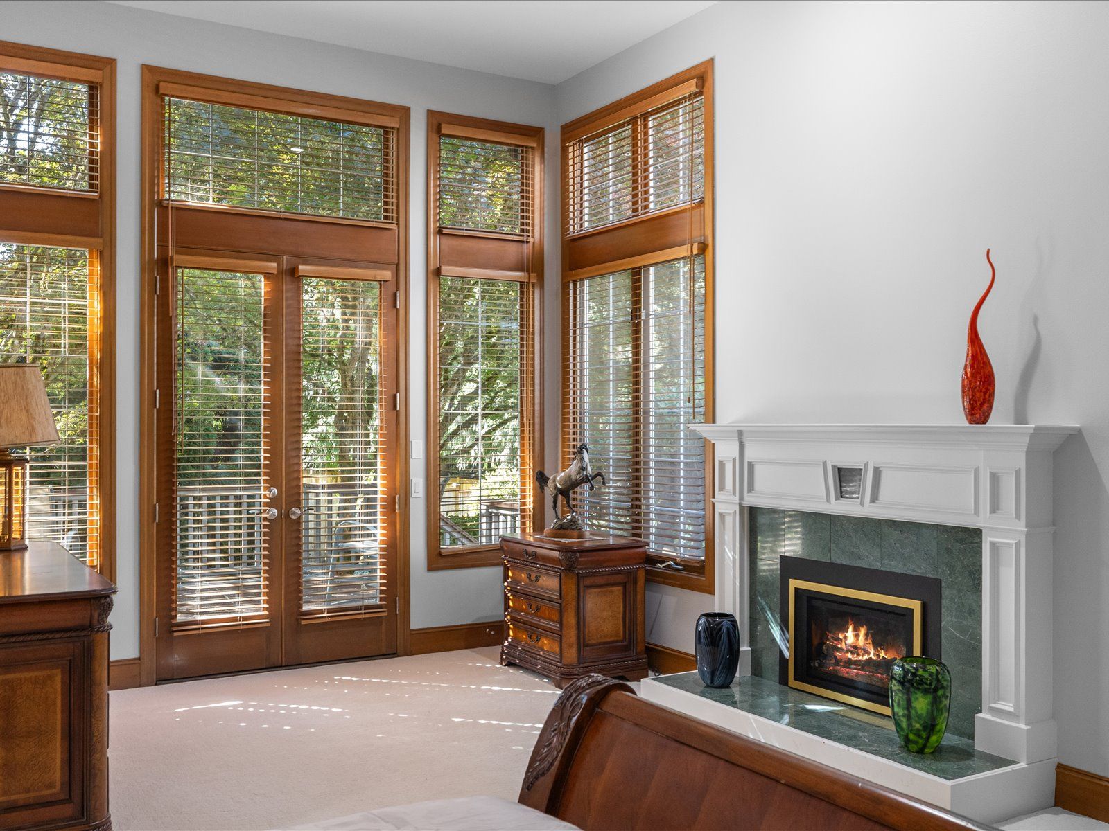 Cozy living room with hardwood floors, large windows overlooking a lake, wood-paneled ceiling, floral sofa, armchairs, wooden coffee table with flowers, and red curtains, in a lakeside cabin or home.