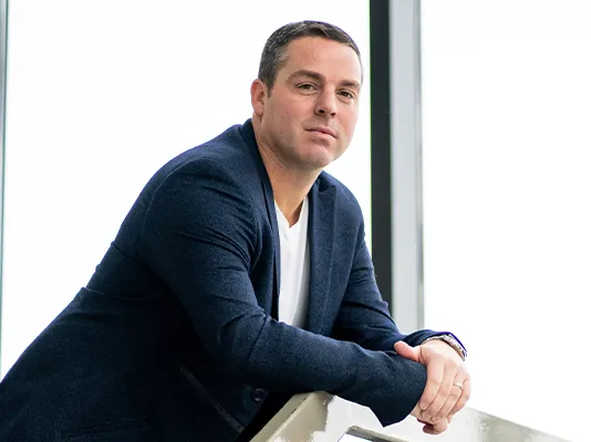 Confident young man in a navy blazer leaning on a railing and looking at the camera near large windows in a modern office setting