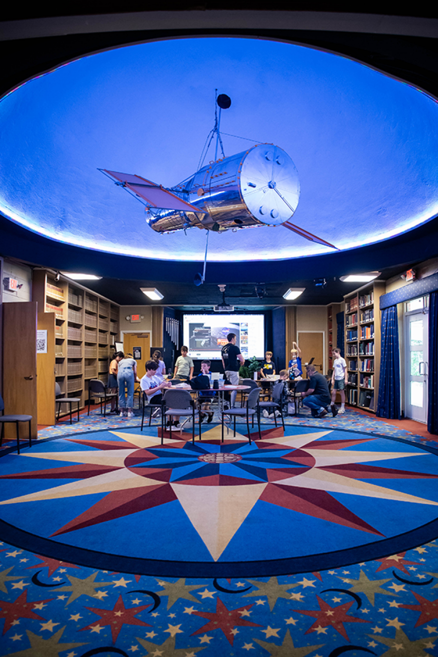 Interior view of a space-themed classroom with a model satellite hanging from the ceiling, students working at tables, and a large digital screen displaying space images in the background.