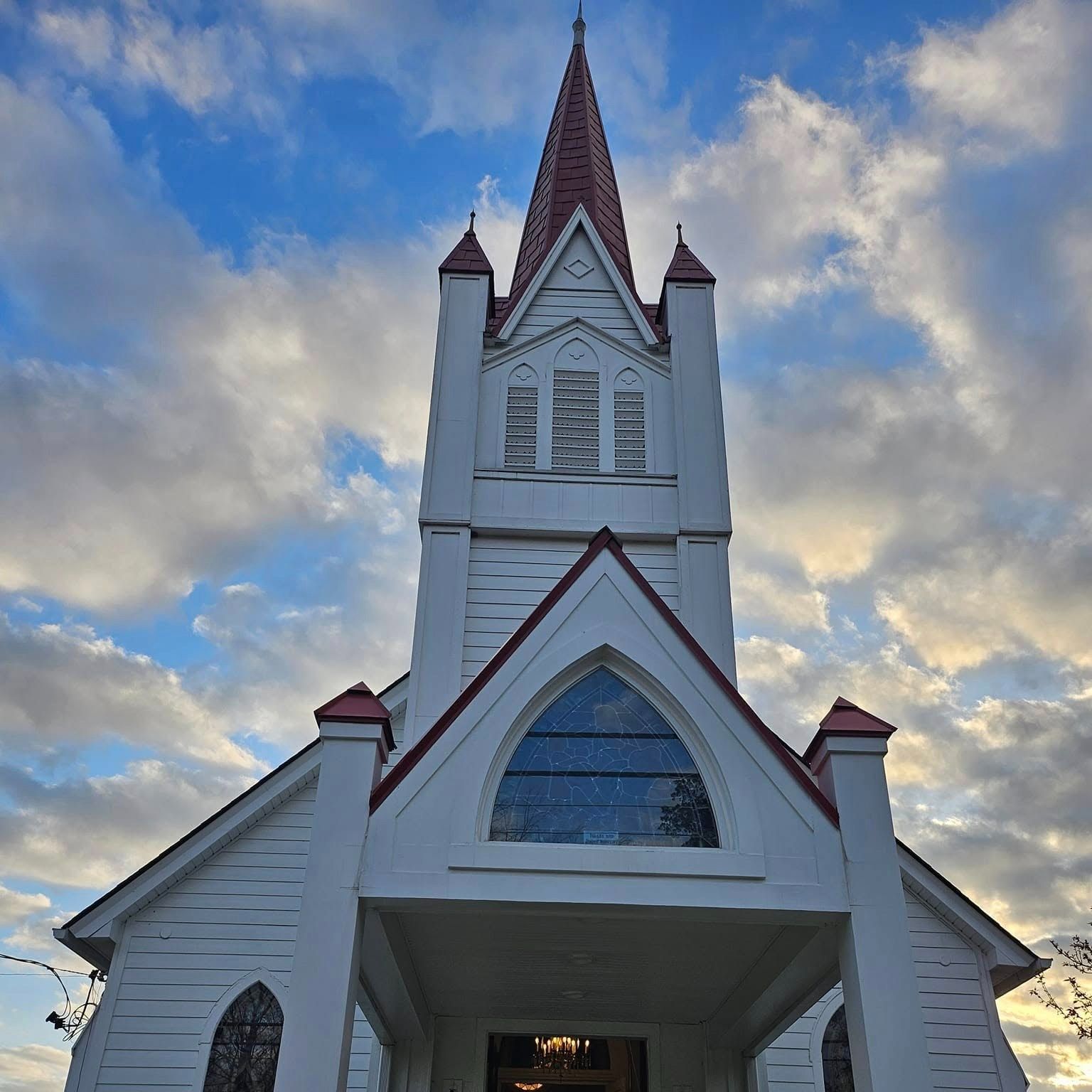 White wooden church with red roof accents and a tall steeple against a partly cloudy sky during daytime