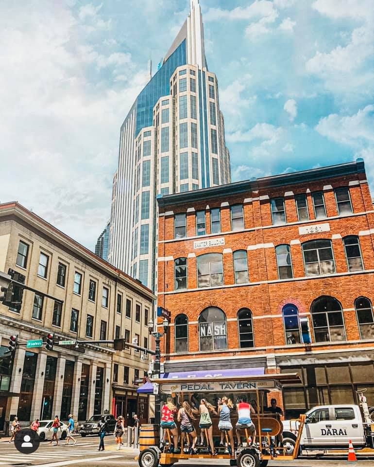 Urban city street scene featuring a modern skyscraper, historic brick building, Pedal Tavern bike bar, and pedestrians under a partly cloudy sky