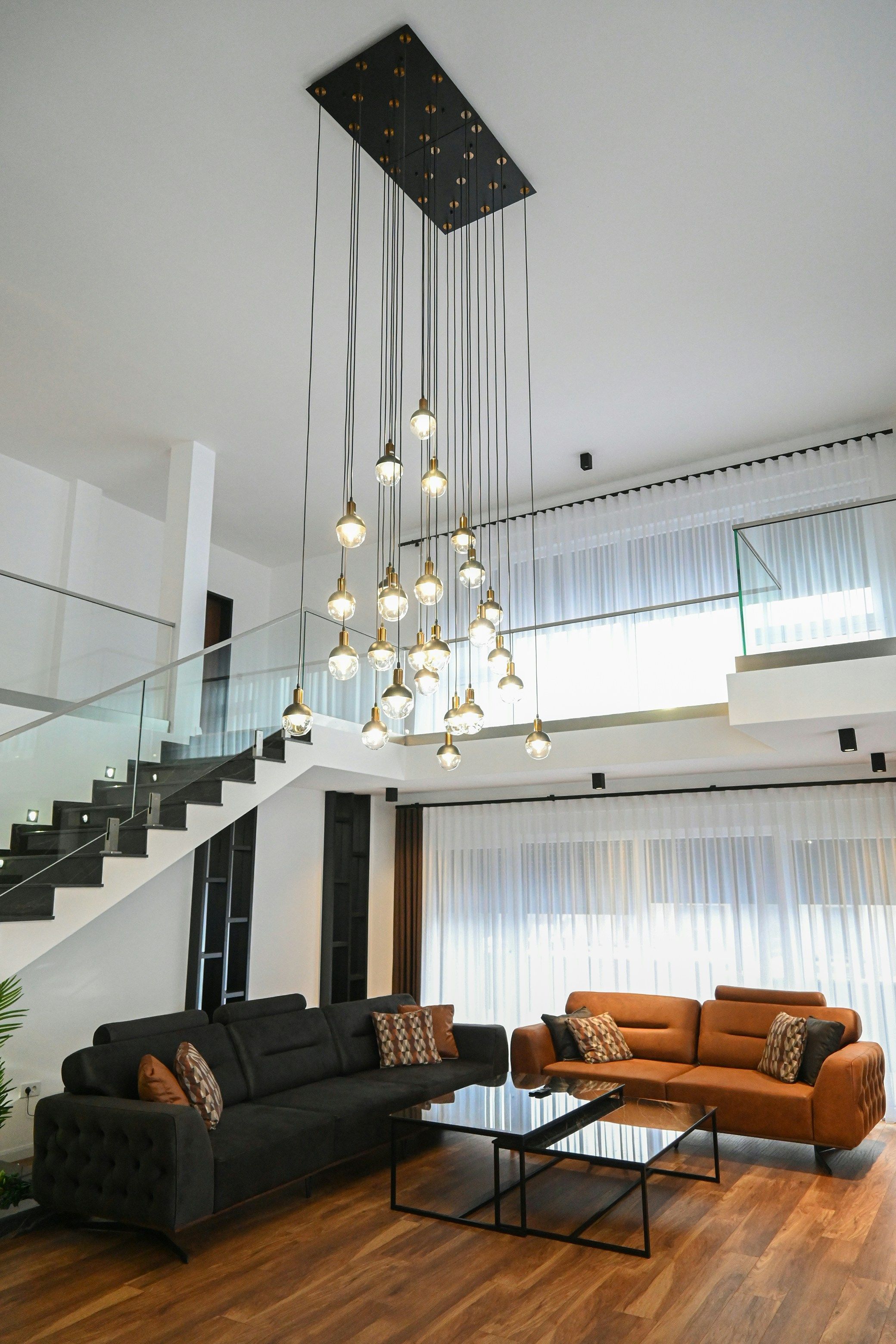 Modern luxury living room with contemporary black and brown sofas, geometric coffee table, large windows with sheer curtains, and a striking chandelier with multiple bulbs hanging from the ceiling in a spacious, stylish home interior.