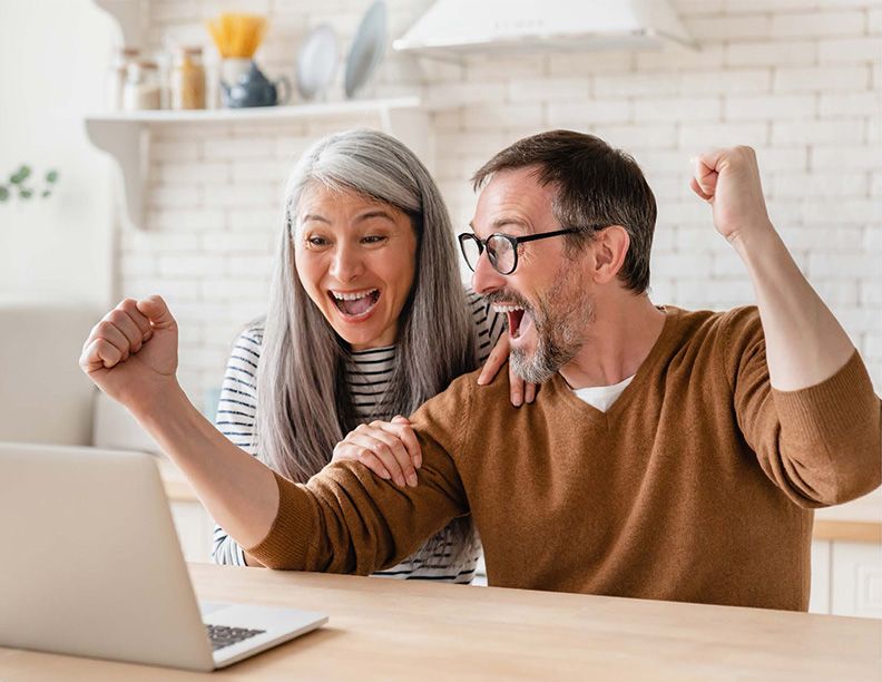 Happy middle-aged couple celebrating success while looking at a laptop in a modern kitchen.