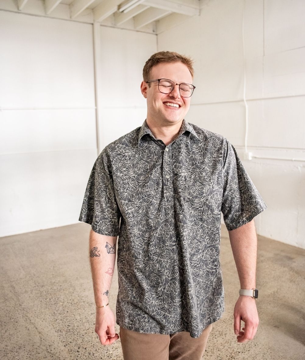 Smiling man wearing a patterned top and glass with a warehouse setting in the background.