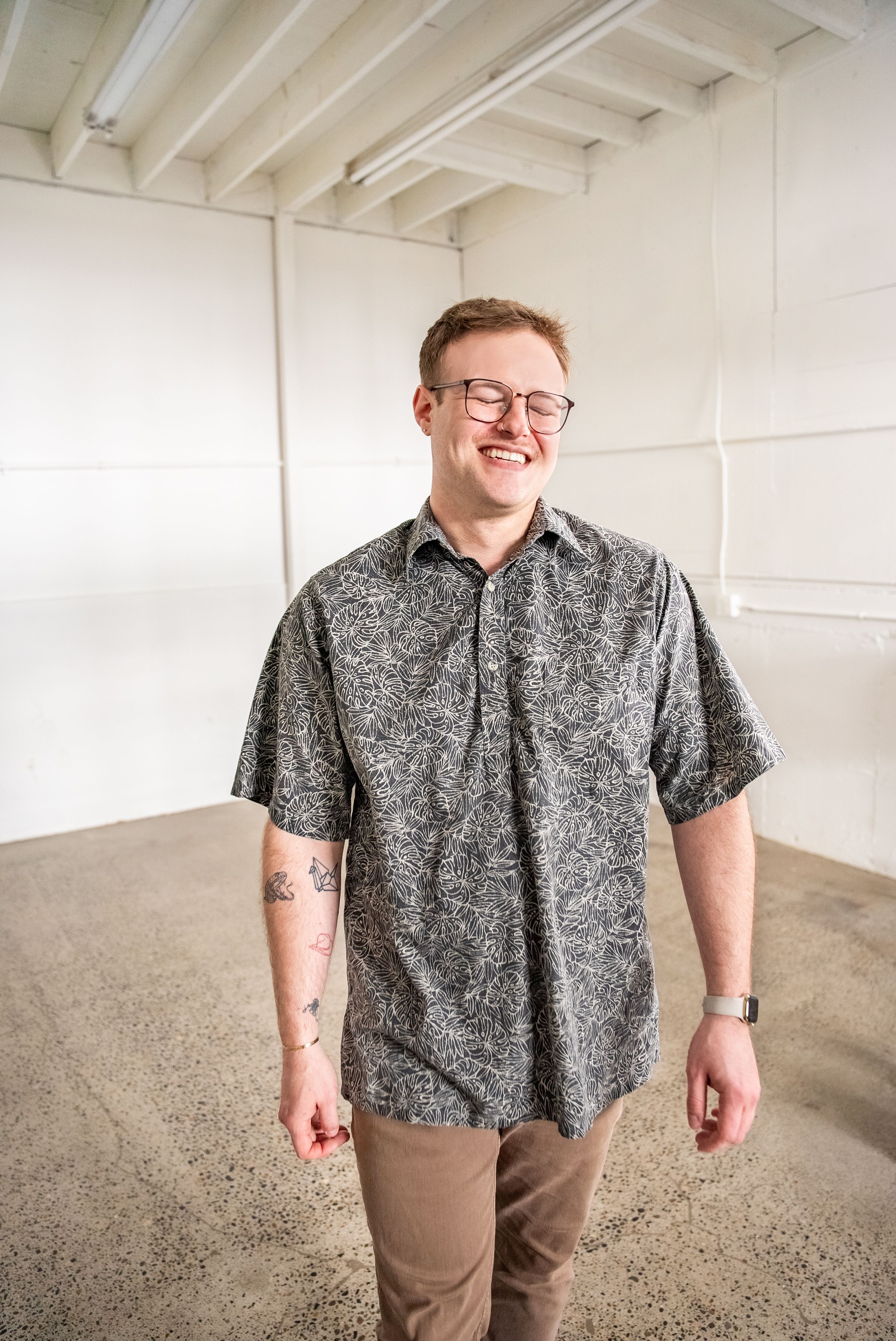 Smiling man wearing a patterned top and glass with a warehouse setting in the background.