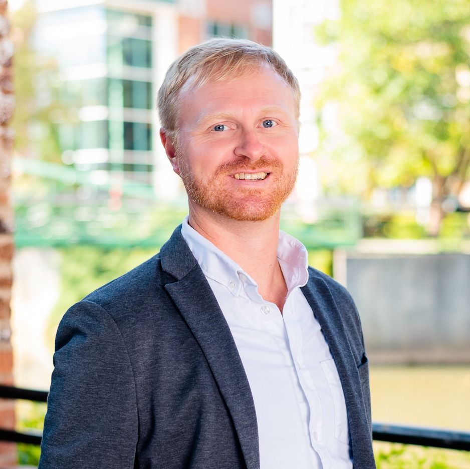 Portrait of a smiling man with dark brown hair and mustache wearing a navy blue button-up shirt against a plain gray background