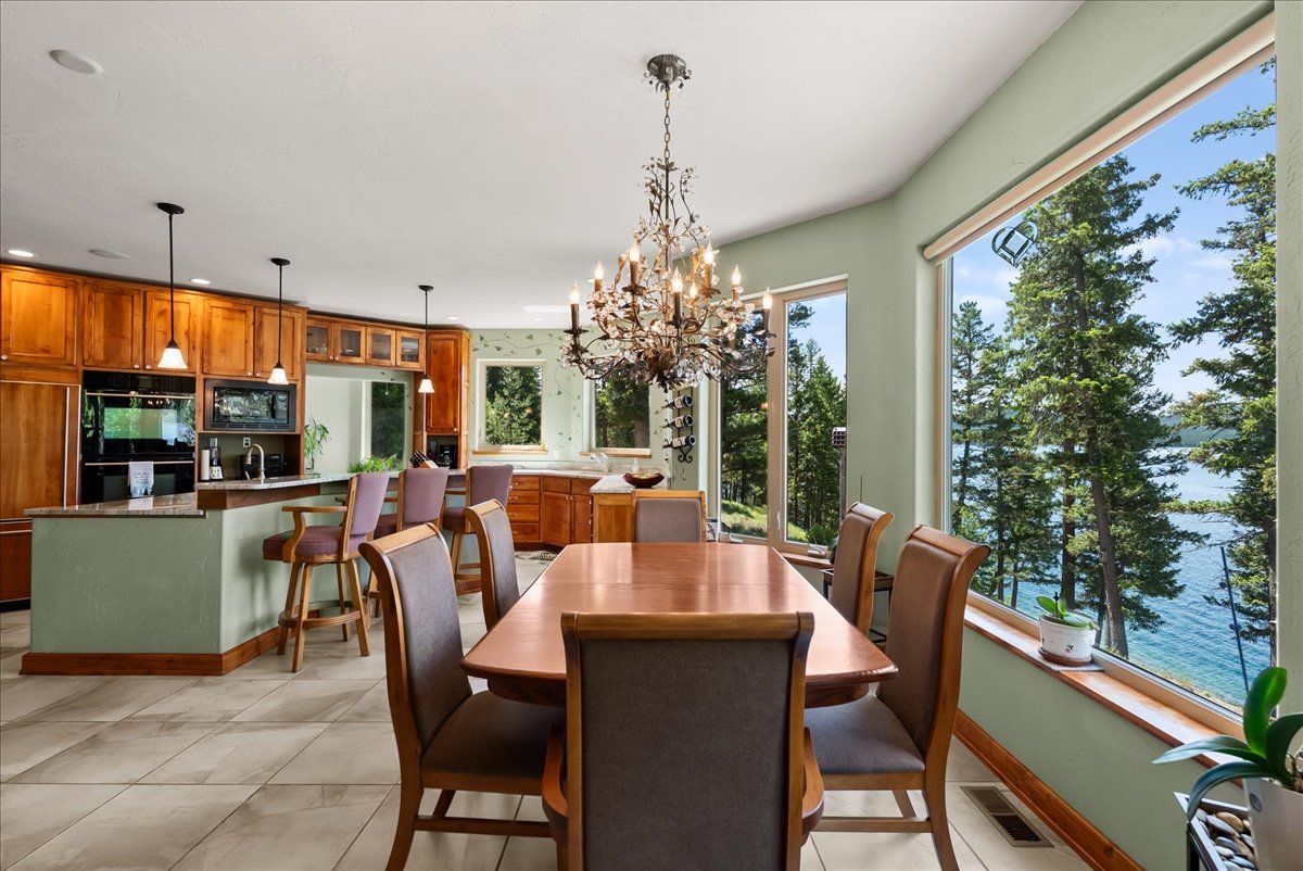 Spacious living room with large windows overlooking a wooded outdoor view, featuring a gray sofa, two armchairs, a colorful area rug, a wooden coffee table, a wood-burning stove, and a modern ceiling fan.