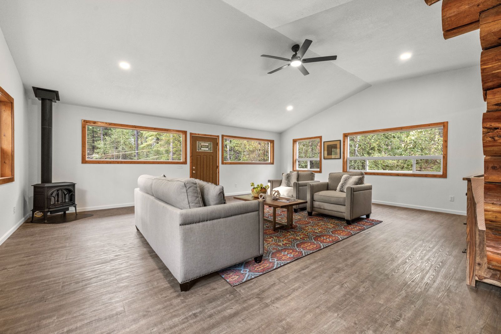 Spacious living room with large windows overlooking a wooded outdoor view, featuring a gray sofa, two armchairs, a colorful area rug, a wooden coffee table, a wood-burning stove, and a modern ceiling fan.