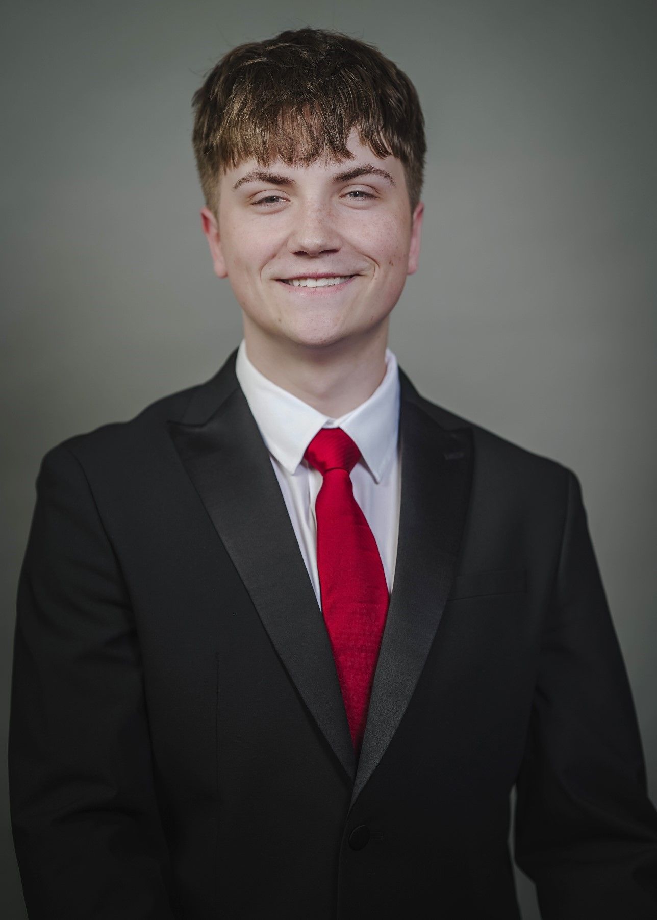 Young businessman in formal black suit, white shirt, and red tie smiling against a plain gray background.
