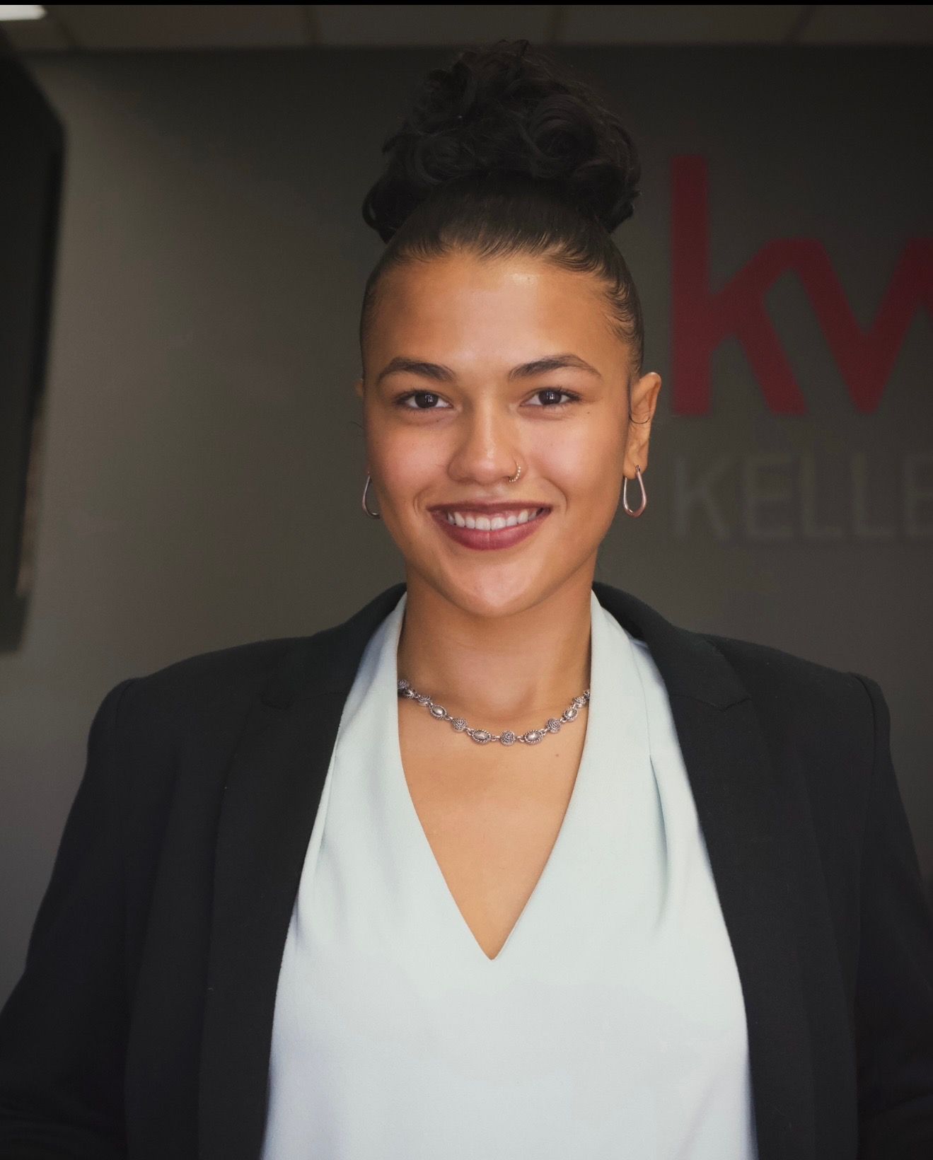 Professional woman smiling with dark hair in a high bun, wearing a black blazer, white blouse, jewelry, and earrings, standing in front of a gray background.