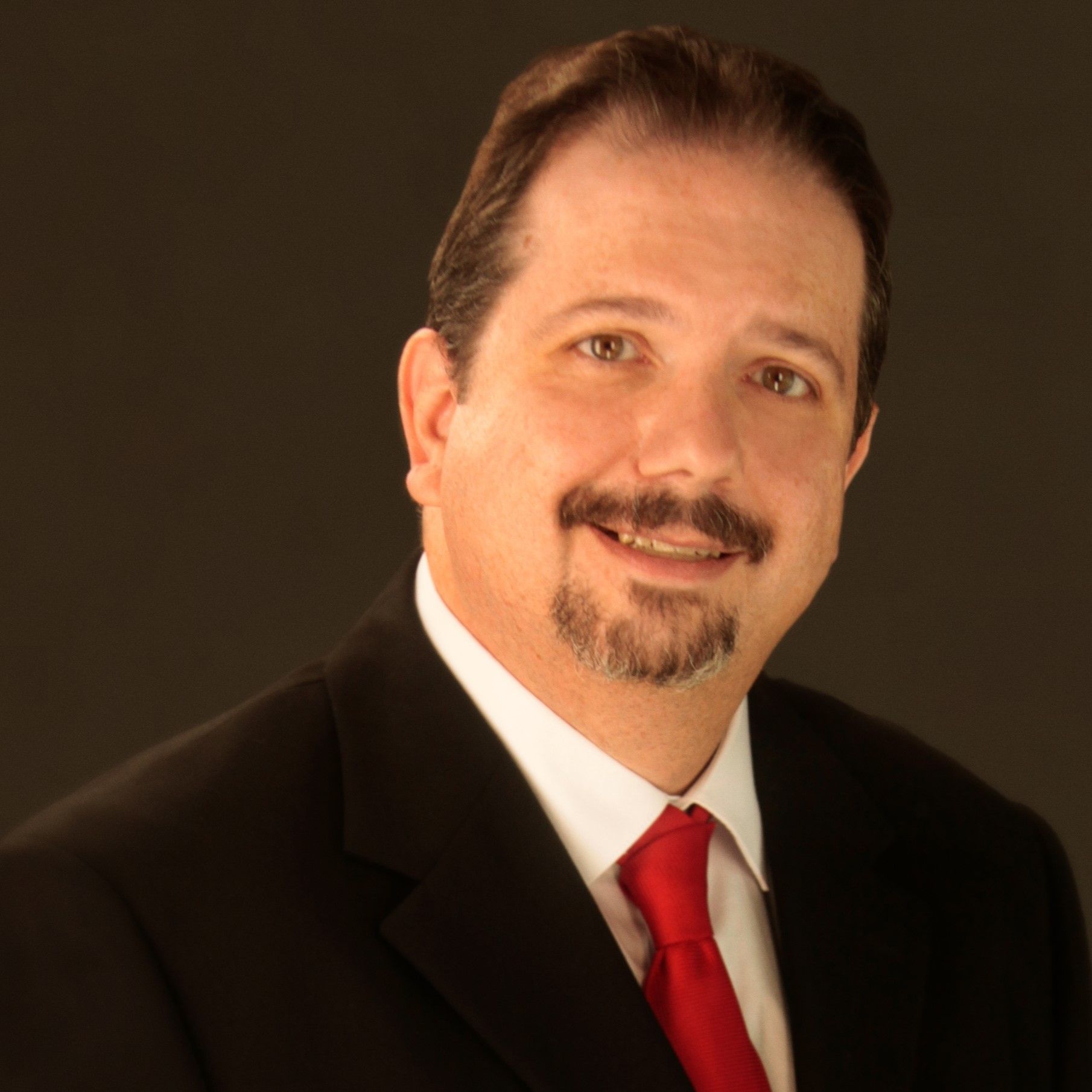 Professional headshot of a middle-aged man with dark hair, styled back, wearing a black suit, white shirt, and red tie, against a dark background.