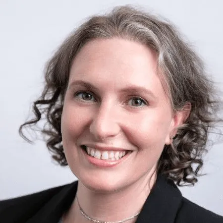 Portrait of a smiling woman with curly gray hair wearing a black blazer and silver necklace on a light gray background